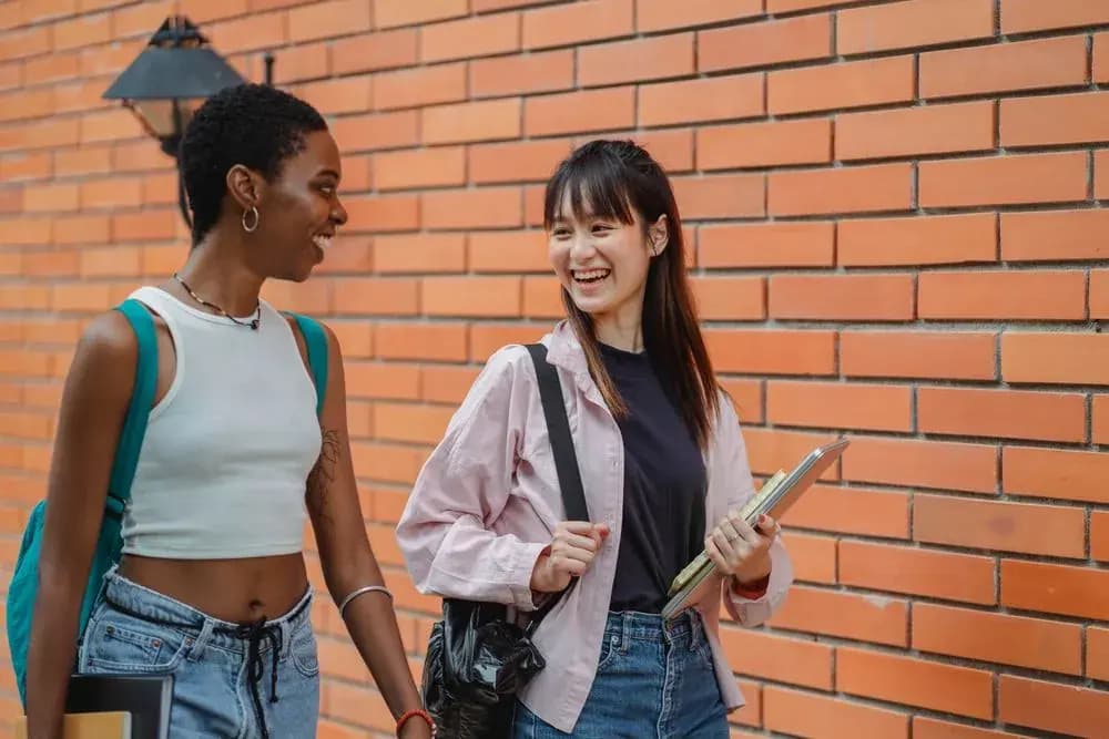 Two smiling students with backpacks and notebooks walking past a brick building